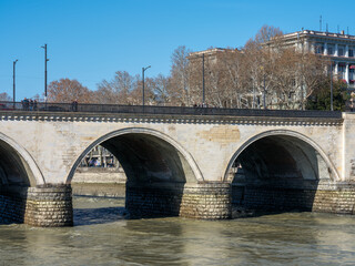 Historical stone bridge with multiple arches spanning across the Mtkvari River in Tbilisi, Georgia, with city buildings and bare trees in the background under a clear blue sky. © Microscope