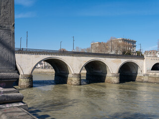Historical stone bridge with multiple arches spanning across the Mtkvari River in Tbilisi, Georgia, with city buildings and bare trees in the background under a clear blue sky. © Microscope