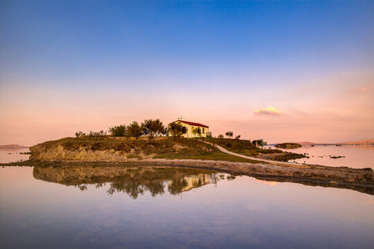 Lemnos Nea Koutali Saint Nicolas islet, Church on rugged islet reflecting in still bay water, featuring clear sky with soft sunset colors. Serene religious landscape