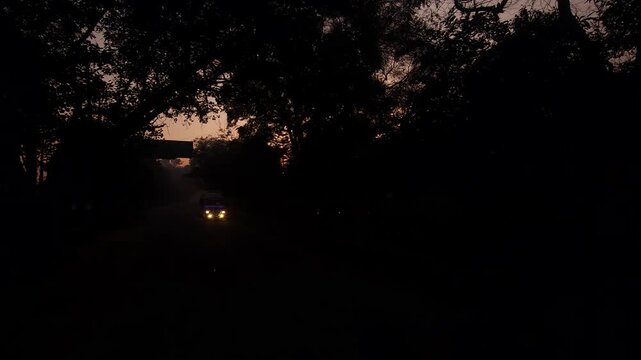 POV shot from a moving vehicle driving on road during sunrise in India. Golden morning light with road side trees and homes and silhouettes. Concept of travel, daily commute, journey