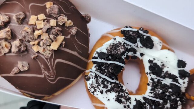 Sweet bakery treats featuring rich icing, cookie crumbs, and decorative drizzle, presented fresh inside a takeaway package for indulgent snacking. Chocolate Glazed Doughnuts With Toppings in Box Close