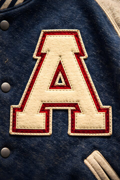 Close-up of a cream-colored felt letter A with red trim, stitched onto a navy blue varsity jacket.