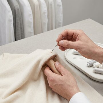 Close up of hands darning a grey wool sweater with a needle and thread using a wooden darning mushroom for garment repair and sustainable fashion maintenance at home.