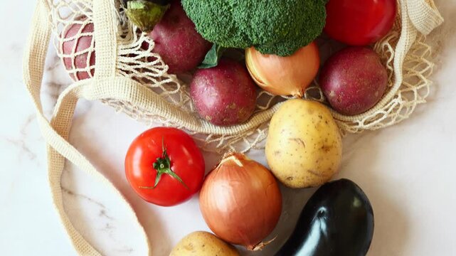 Fresh eggplants, red and yellow potatoes, onions, and red tomatoes rest on a white marble surface next to a reusable mesh bag.