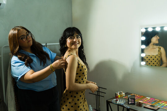 Professional stylist supporting young woman backstage, adjusting her dress before a fashion event with makeup table nearby