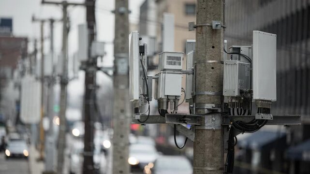 Cluster of small microcell devices aligned on utility poles overhead in a dense urban passage foremost unit sharply captured against a blurred street scene.