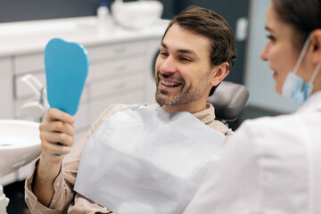 Happy male patient smiling while looking in mirror during dental appointment at clinic, being satisfied with result of procedure