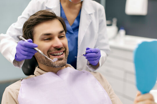 Man patient looking at mirror while dentist pointing at his teeth with tools and showing result of treatment