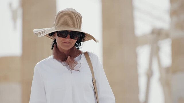 Fashionable woman wearing sun hat and sunglasses walks near ancient architectural ruins of Parthenon on sunny day in Athens, Greece