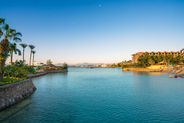Fototapeta premium A beautiful morning view of a turquoise lagoon at a luxury resort in Hurghada, Egypt, featuring palm trees, hotel buildings, and clear blue skies.