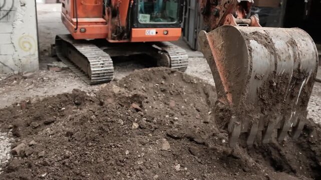 Close-up video of a heavy-duty orange hydraulic excavator bucket digging a deep narrow trench in dark, moist soil at an urban construction site. The video shows the precise mechanical movement.