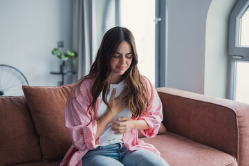 Young woman sits on a living room couch clutching her chest in visible discomfort, suggesting...
