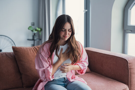 Young woman sits on a living room couch clutching her chest in visible discomfort, suggesting heartburn, indigestion or anxiety-related chest pain in a domestic setting.