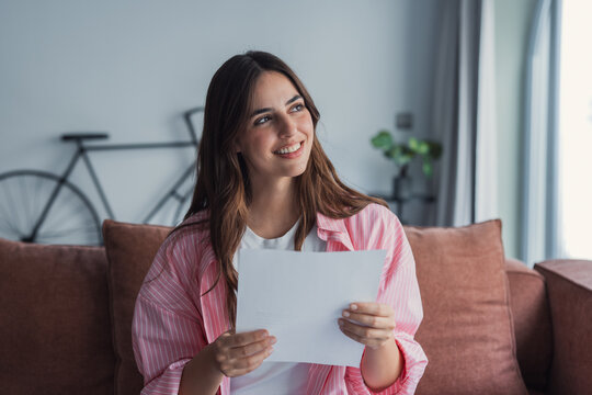 Smiling young woman reading a letter at home on a couch, relaxed lifestyle and happy reaction to good news