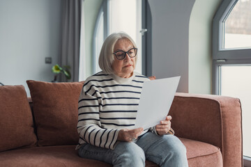 Obraz premium Elderly woman reading paperwork on sofa at home, senior with glasses reviewing a letter in a quiet living room