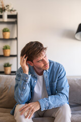 Obraz premium A young man in a denim shirt sits on a sofa at home, rubbing his head and looking pensive. The candid portrait conveys stress, worry and quiet contemplation in a relaxed living room setting.