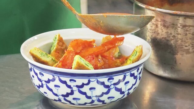 Ladle pouring hot Thai sour curry (Kaeng Som) with shrimps and acacia omelet into a traditional ceramic bowl.