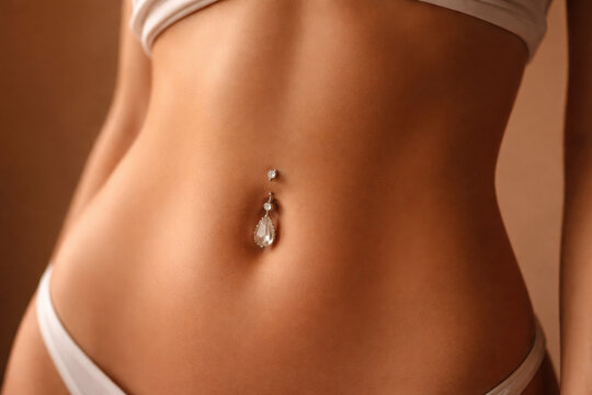 Close-up of European woman abdomen with belly button piercing on brown background, minimal aesthetic body jewelry and lifestyle concept