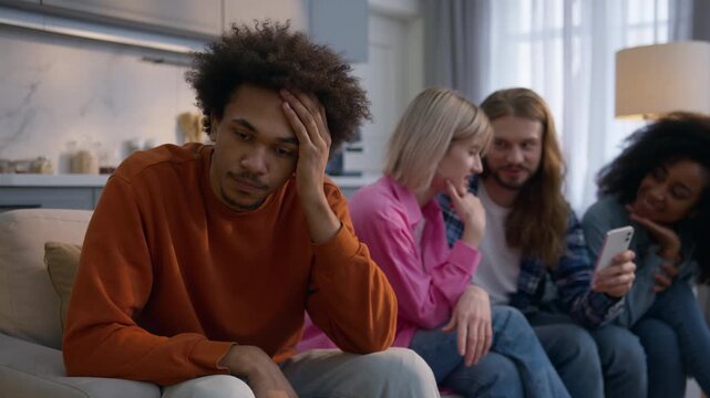 Stressed sad African American man sitting home frustrated male introvert headache ignoring friends while multiethnic people chatting mobile phone cellphone together social isolation gadget addiction