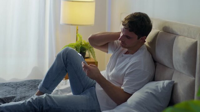 Sleepy tired Caucasian man lying bed bedroom home yawning using mobile phone scrolling social media laziness European bored male guy hold smartphone chatting online technology morning awakening indoor