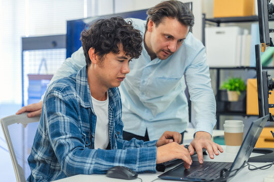 collaborative AI development team in modern tech office: young male developer coding on laptop while senior colleague leans over discussing code and data visualizations on multiple monitors