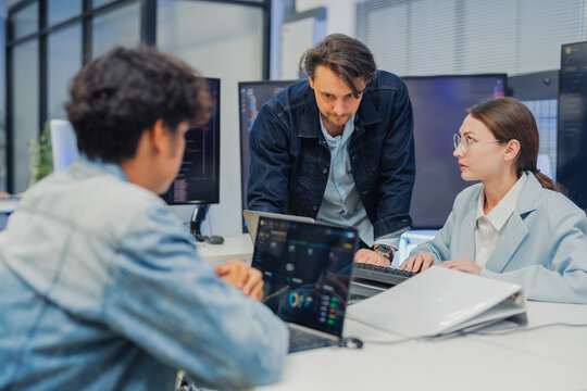 collaborative AI development team in modern tech office: young male developer coding on laptop, female team leader in light blue blazer pointing at code and neural network visualization 