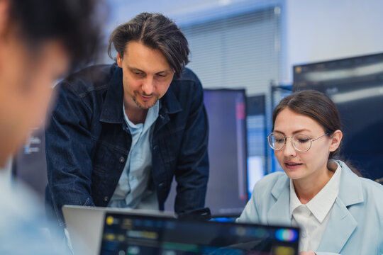 collaborative AI development team in modern tech office: young male developer coding on laptop, female team leader in light blue blazer pointing at code and neural network visualization 