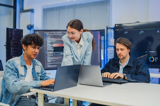 collaborative AI development team in modern tech office: young male developer coding on laptop, female team leader in light blue blazer pointing at code and neural network visualization 