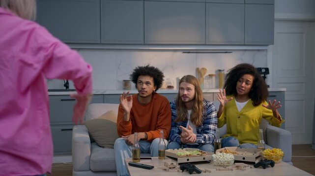 Multiracial friends playing guessing game at home sofa Caucasian woman showing pantomime explaining word fun diverse African women men teenagers laughing trying to guess charades together indoor party