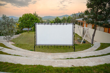 Outdoor cinema screen setup on a grassy area with mountain view and beautiful sunset sky. Concept of relaxation, outdoor entertainment, camping lifestyle, and nature evening activity.