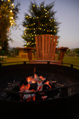 A cozy outdoor scene with a burning campfire in front of a wooden chair and a Christmas tree decorated with string lights. Perfect winter evening atmosphere for relaxation, camping