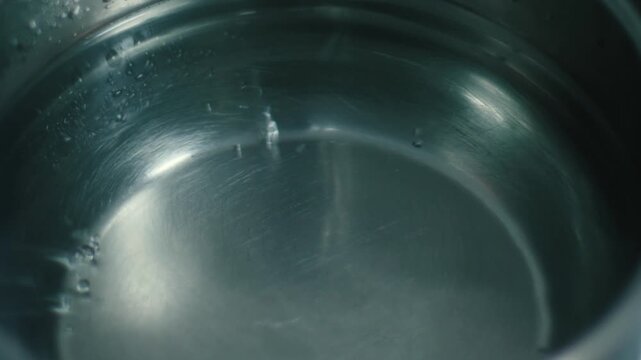 Close up of water starting to boil in a stainless steel pot. Air bubbles rise from the bottom of the metal cookware during the heating process.