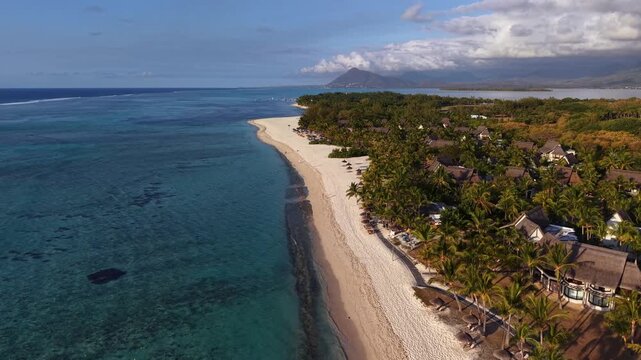Aerial view shows a tropical beach with clear turquoise waters and white sands. Lush greenery and resort-style huts line the shore. Few people relax under umbrellas near the water