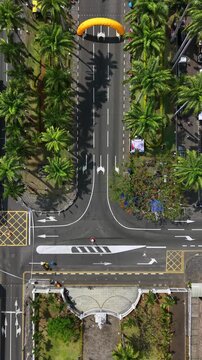 Aerial view of a vibrant tropical street lined with palm trees. Bright yellow parasail glides over the road, casting shadows on asphalt. Traffic moves calmly beneath the canopy of lush green palms