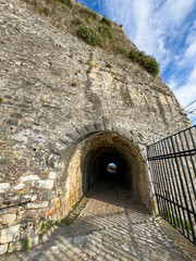 Fototapeta premium Stone tunnel entrance at the New Fortress in Corfu, Greece. Cobblestone path, iron gate, blue sky, sunny day