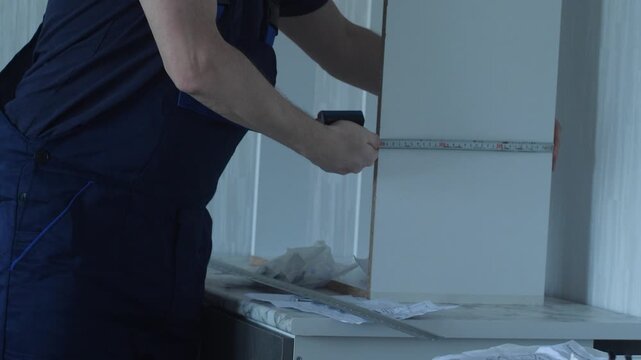 Professional furniture assembler measuring kitchen cabinet parts with a tape measure. Man in blue overalls studying drawings and checking furniture dimensions