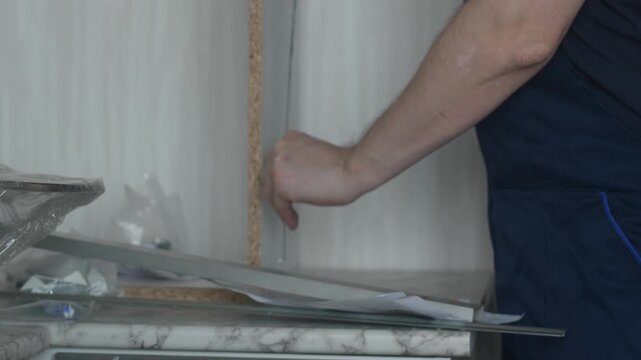 Professional furniture assembler measuring kitchen cabinet parts with a tape measure. Man in blue overalls studying drawings and checking furniture dimensions.