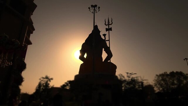 Silhouette of Lord Shiva statue with trishul near a temple during golden sunset in India. Spiritual Hindu scene with devotees, dramatic sky, Chaar Dham Temple in Vrindavan, Uttar Pradesh.