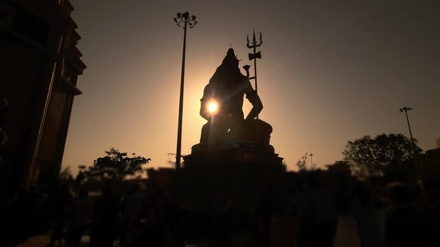 Silhouette of Lord Shiva statue with trishul near a temple during golden sunset in India. Spiritual Hindu scene with devotees, dramatic sky, Chaar Dham Temple in Vrindavan, Uttar Pradesh.