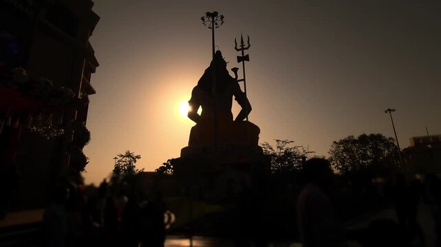 Silhouette of Lord Shiva statue with trishul near a temple during golden sunset in India. Spiritual Hindu scene with devotees, dramatic sky, Chaar Dham Temple in Vrindavan, Uttar Pradesh.