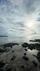 Fototapeta premium Serene Tropical Coastline with Dark Volcanic Rocks and Calm Ocean Under a Cloudy Sky