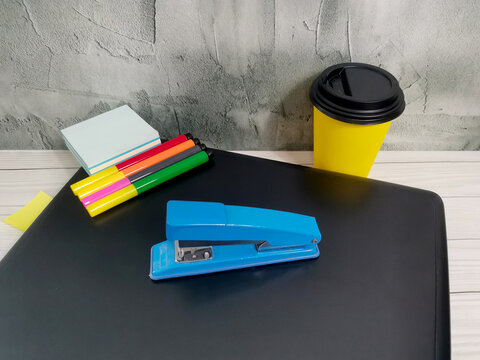 Blue stapler with colorful markers, sticky notes and yellow coffee cup on black desk. Gray textured wall background. Office supplies for work, study, business organization workspace