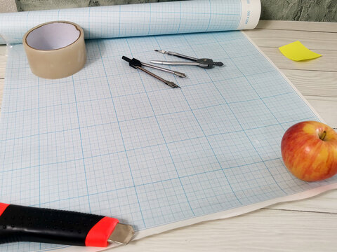 Blue grid paper roll with drawing compass, tape, utility knife and apple on white wooden desk. Drafting tools for architecture, engineering, design, technical drawing workspace