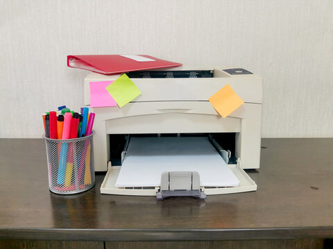 Beige printer with red folder and colorful sticky notes on dark wooden desk. Mesh holder with markers beside. Home office, business documentation, remote work workspace environment