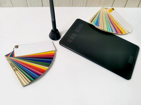 Black graphics tablet with colorful swatches and stylus holder on white desk. Design workspace for creative projects, digital art, color selection