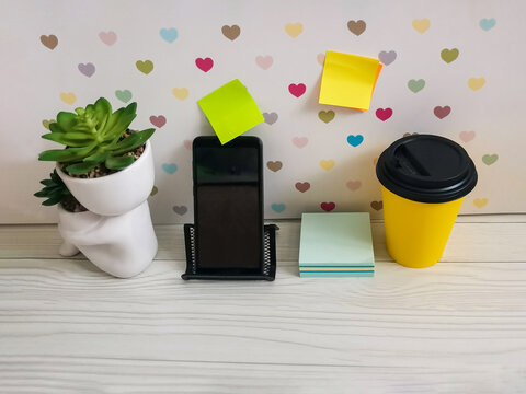 Yellow coffee cup, smartphone on stand and sticky notes on white wooden desk with heart pattern wallpaper. succulent plant. Workspace for freelance, study, business, remote work