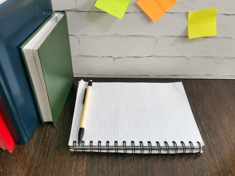 Open spiral notebook with grid paper and pen on dark wooden desk. Books and colorful sticky notes on white brick wall background. Study, work, planning, education, note taking workspace
