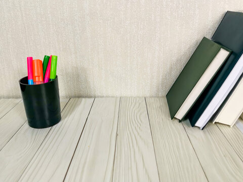 Books with colorful highlighters in black holder on white wooden desk. Textured beige wall background. Study, work, office supplies, education, organization workspace with copyspace