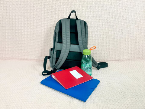 Gray backpack with red notebook, blue folder for documents and green water bottle on sofa. School supplies, student essentials, education, study accessories