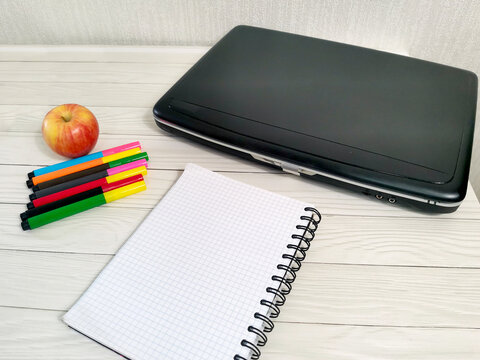 Closed black laptop with spiral notebook, colorful markers and fresh apple on white wooden desk. Study supplies for remote work, education, business workspace setup.
